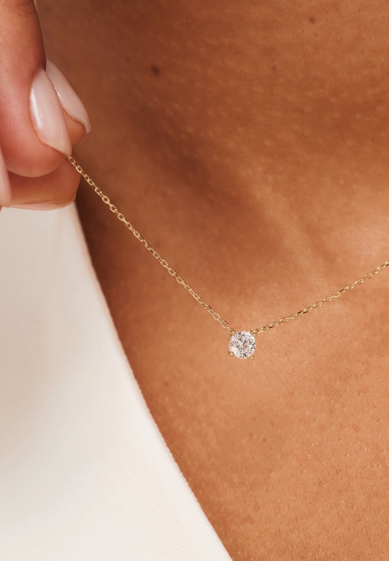 Collier en or avec un petit pendentif en diamant rond à six griffes, posé sur un fond de teint. Design simple et élégant.