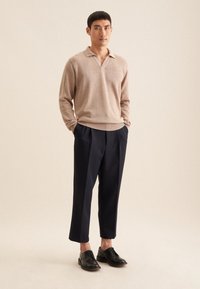 Beige gestrickter Pullover mit Kragen, marineblaue maßgeschneiderte Hose und schwarze Lederschuhe. Person steht mit den Händen in den Taschen vor einem neutralen Hintergrund.