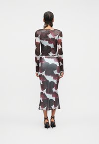 Γυναίκα με πλεκτά μαλλιά που φορεί εφαρμοστό φόρεμα μακρυμάνικο μέχρι τους αστραγάλους με μεγάλα σκούρα λουλουδάτα μοτίβα και μαύρες ψηλοτάκουνες σανδάλες.