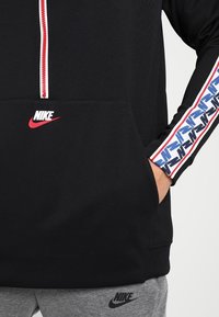 Svart huvtröja med vit dragkedja, rött Nike-logotyp på bröstet och mönstrade blå och röda accenter på ärmarna. Lättviktigt tyg.