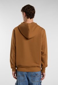 Persona con capelli ricci, che indossa una felpa marrone e pantaloni cargo in denim blu, di spalle contro uno sfondo chiaro e semplice.