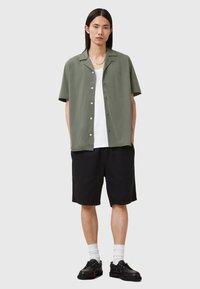 Kortermet olivengrønn skjorte med knapper, kombinert med svarte shorts, hvit skjorte under, hvite sokker og svarte sko.