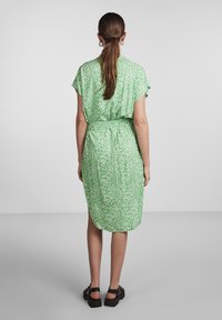 Grönblommig klänning med korta ärmar, midjebälte, knälång fåll och sidosprund. Buren med kraftiga svarta sandaler.