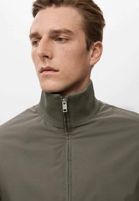 Chaqueta verde oliva con cremallera, de cuello alto, que presenta una tela suave y una cremallera metálica en la parte delantera. Diseño minimalista sin patrones.