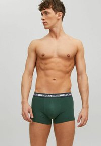 Fit man with short curly hair wearing green Jack & Jones boxer briefs, looking to his left against a plain light gray background.