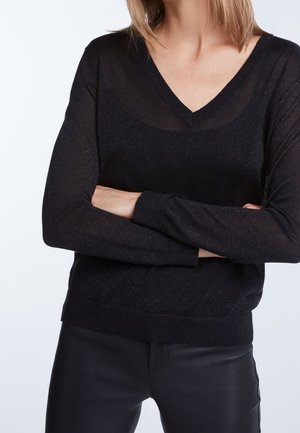 Mujer con los brazos cruzados usando un suéter negro de cuello en V transparente y pantalones de cuero negros, frente a un fondo blanco.