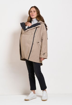 Kvinde iført en beige bærejakke, der holder en sovende baby tæt ved sit bryst, iført sorte bukser og hvide sneakers.