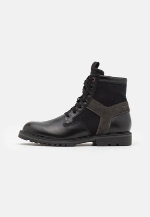 Sort ankelstøvle i læder og stof med snørebånd, robust sål og ruskindskrydsdetalje på siden mod en hvid baggrund.