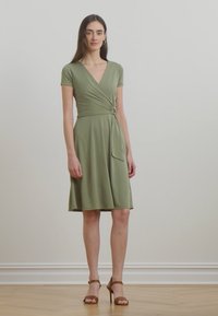 Vestido envolvente verde-azeitona com mangas curtas, tecido suave, saia até ao joelho e detalhe de laço lateral. Notável pelo seu design simples e pelo ajuste.