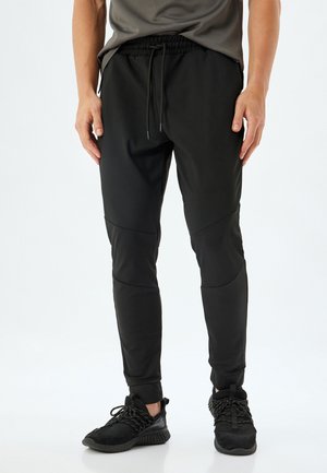 Zwarte sportbroek met een elastische tailleband, een trekkoord en een tapered fit. Heeft paneeldetails en geribbelde manchetten. Gecombineerd met zwarte schoenen.