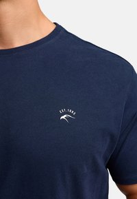 Granatowa bawełniana koszulka z okrągłym dekoltem, z małym białym logo i napisem "EST.1993" na lewej klatce piersiowej. Gładka tekstura.