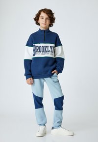 Marinblå sweatshirt med vita och gröna ränder, med texten "BROOKLYN". Ljusblå byxor med marinblå lappar, tillsammans med vita sneakers.