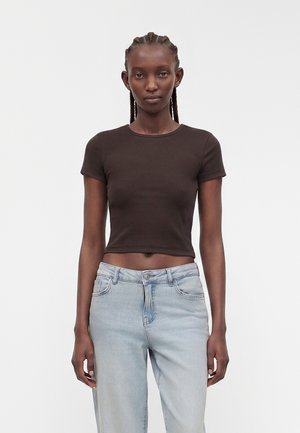 Slank person med fletninger, iført en tætsiddende sort kortærmet crop top og lyseblå højttaljede jeans, står mod en ensfarvet baggrund.