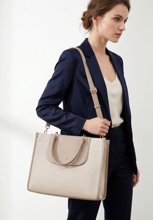 Jonge vrouw in marineblauw pak houdt een beige gestructureerde tote bag met dubbele handvatten en een schouderriem aan haar rechterarm.