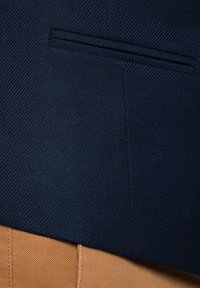 Granatowy materiał w jodełkę o strukturze z jedną kieszenią na piersi, w połączeniu z brązowymi spodniami o gładkiej, tkanej fakturze.