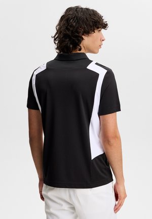 Mann mit lockigem dunklem Haar, der ein schwarzes Poloshirt mit weißen geometrischen Einsätzen an den Seiten und weiße Hosen trägt, von hinten vor einem schlichten Hintergrund.