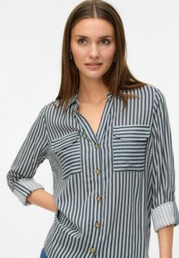 Jeune femme portant une chemise boutonnée à rayures verticales noires et blanches avec des manches retroussées, se tenant devant un fond blanc uni.