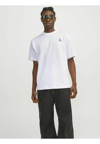 T-shirt bianca in cotone con maniche corte e un logo a triangolo viola sul lato sinistro del petto, abbinata a pantaloni neri a gamba larga.