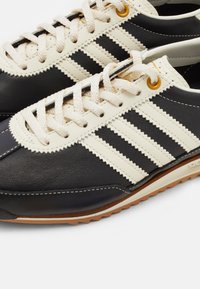Black and cream athletic shoes made of leather, featuring three cream stripes, a textured sole, and round laces. Gold eyelets accent the design.