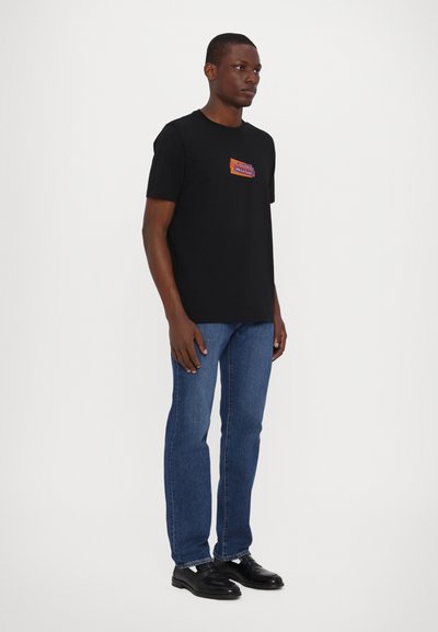 Czarny bawełniany t-shirt z okrągłym dekoltem z kolorową graficzną naszywką na klatce piersiowej, w zestawieniu z niebieskimi dżinsami i czarnymi butami. Gładka tekstura.