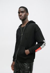 Mlađi muškarac u crnoj zip-up dukserici s bijelim i crvenim natpisom na rukavu i crnim kožnim hlačama s patentnim zatvaračima.