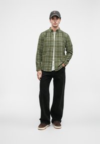 Grønn rutete flanellskjorte over en lys t-skjorte, kombinert med svarte vide bukser og brune joggesko. Tilbehør inkluderer en mørk caps.