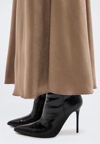 Bottes noires à talons aiguilles et bouts pointus, portées sous une longue jupe fluide beige à la texture lisse.