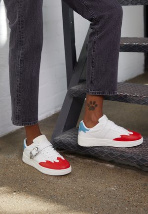 Person iført hvide sneakers med røde og blå accenter, stående på en metaltrappe med en pote-tatovering synlig på deres højre ankel.