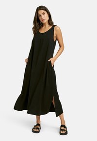 Schwarzes ärmelloses Maxikleid mit Seitentaschen, lockerer Passform, Schlitzen am Saum und glatter Textur. Trägt schwarze Sandalen.
