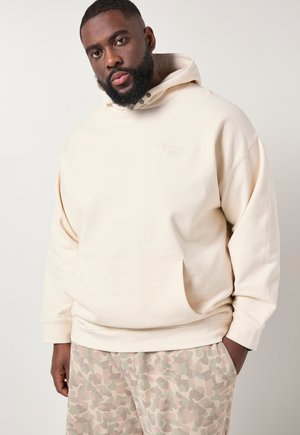 Mann mit Bart, der einen cremefarbenen Hoodie und beige Tarnhosen trägt, steht mit einer Hand in der Tasche vor einem einfarbigen Hintergrund.