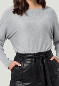 Pull tricot gris clair avec des épaules tombantes et des manches longues, associé à un pantalon en simili cuir noir taille haute avec un détail de ceinture nouée.