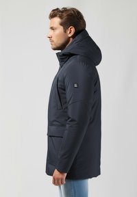 Navy spalvos izoliuota striukė su kapišonu, turinti užtrauktuką priekyje, šonines kišenes ir tekstūruotą medžiagą šilumai. Papuošta logotipu ant rankovės.