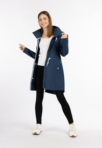 Giacca impermeabile blu navy con colletto alto, chiusura frontale a zip e regolatori a cordino. Presenta due tasche laterali e dettagli bianchi, indossata sopra leggings neri.