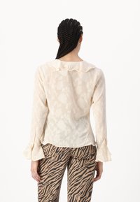 Helle cremefarbene Bluse mit floralem Muster, Rüschkragen und Glockenärmeln; kombiniert mit braun- und schwarzgestreiften Hosen.