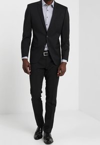 Musta puku, jossa on räätälöity istuvuus, yksirivinen takki, harmaa nappipaita ja kiillotetut mustat kengät.