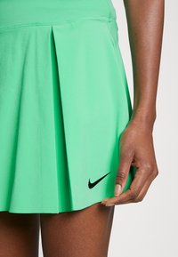 Green pleated tennis skirt made of lightweight fabric, featuring a black Nike logo at the hem and smooth, streamlined design.