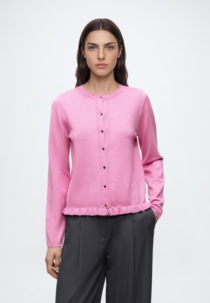 Femme aux cheveux foncés portant un pull rose à boutons et un pantalon ample gris foncé, debout devant un fond uni clair.