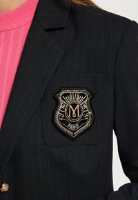 Blazer de rayas navy con parche de cresta negra bordado en oro que dice "M Paris", usado sobre una camiseta de punto acanalado de color rosa brillante.