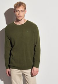 Pull à manches longues côtelé vert avec un col rond et un petit logo sur la poitrine, associé à un pantalon de couleur claire.