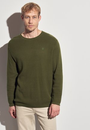 Groene geribbelde trui met lange mouwen en een ronde hals, voorzien van een klein logo op de borst, gecombineerd met lichte broek.