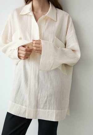 Osoba regulująca mankiety oversize’owej, teksturowanej kremowej koszuli z długim rękawem w połączeniu z czarnymi spodniami na jednolitym białym tle.