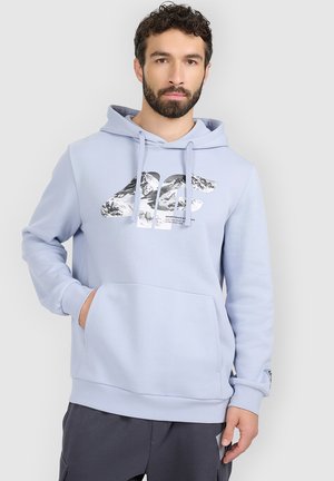 Mężczyzna z brodą, ubrany w jasnoniebieską bluzę z kapturem z grafiką gór i czarne spodnie, stoi z jedną ręką w kieszeni na jednolitym tle.