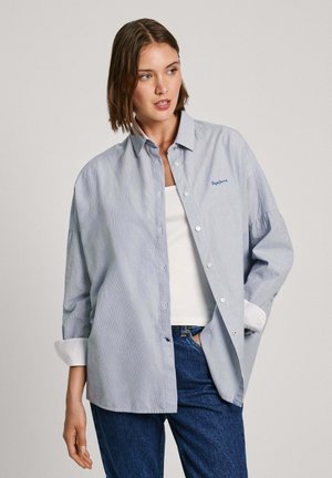 Helleblaue gestreifte Baumwollhemd mit langen Ärmeln, die an den Bündchen aufgerollt sind. Darunter trägt er ein weißes Unterhemd und blaue Jeans.