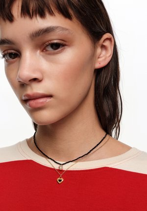 Persona joven con cabello castaño que lleva una camiseta roja y beige y dos collares, uno negro con cuentas y una perla, y otro dorado con un colgante en forma de corazón.