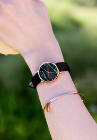 Okrągły zegarek z czarnym paskiem i zieloną tarczą w paski. Zauważalne różowe złote akcenty oraz świecące znaczniki. Widoczna jest również prosta metalowa bransoleta.