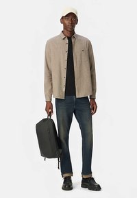 Langarm geruit overhemd in beige, zwart T-shirt, donkere spijkerbroek, zwarte rugzak en zwarte schoenen, gecombineerd met een lichtgekleurde pet.