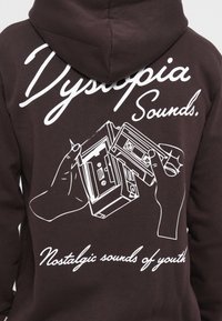 Mørkebrun hoodie med hvid grafisk design af hænder, der holder et kassettebånd og en boombox. Teksten lyder "Dystopia Sounds. Nostalgiske lyde fra ungdommen."