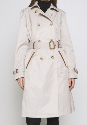 Kobieta w beżowym płaszczu trencz z podwójnym rzędem guzików, brązowym paskiem, zapinanymi na guziki mankietami i ukośnymi kieszeniami, nosząca czarny golf i spodnie.