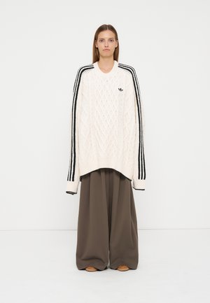 Giovane donna che indossa un maglione bianco oversize lavorato a trecce con strisce nere sulle maniche e pantaloni larghi marroni e morbidi.