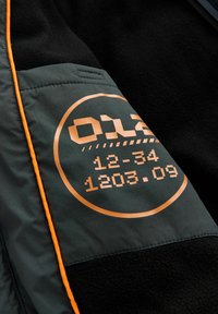 Mørk grøn jakke med orange detaljer. Har et stort kobberfarvet rundt mærke med tal og symboler, samt et tekstureret sort stof.